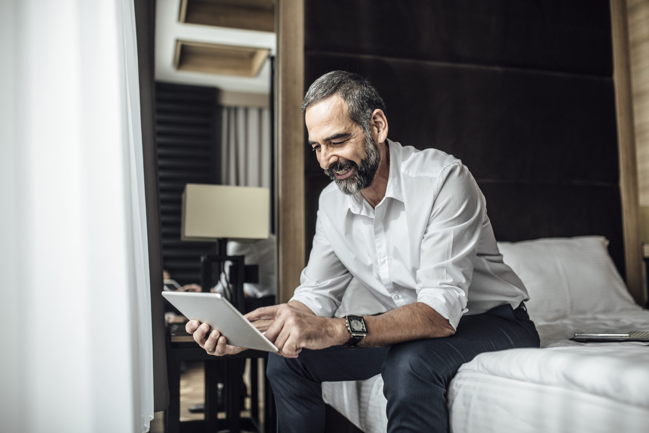 Handsome Caucasian middle-aged businessman sitting on a bed and reading on tablet.